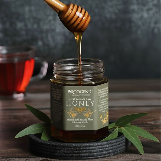 Jar of Roogenic Australian Raw Honey with a wooden honey dipper and leaves on a wooden surface.